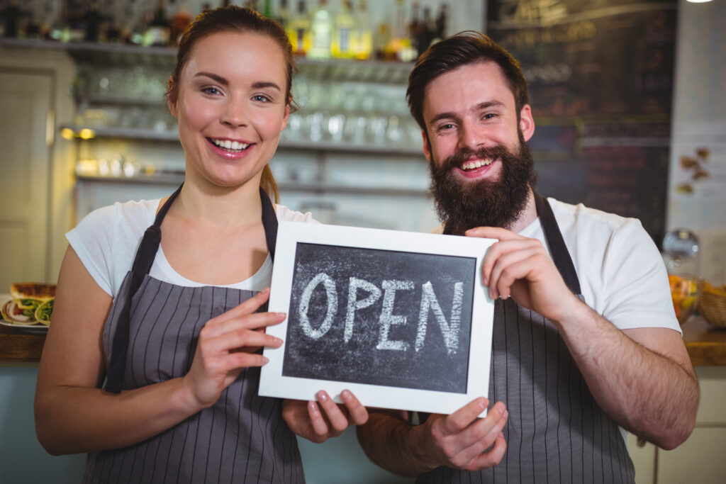 smiling waitress and waiter standing with open sign board in cafe